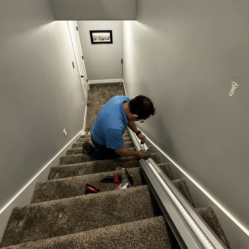An installer fitting a stair lift rail partway up a carpeted basement staircase