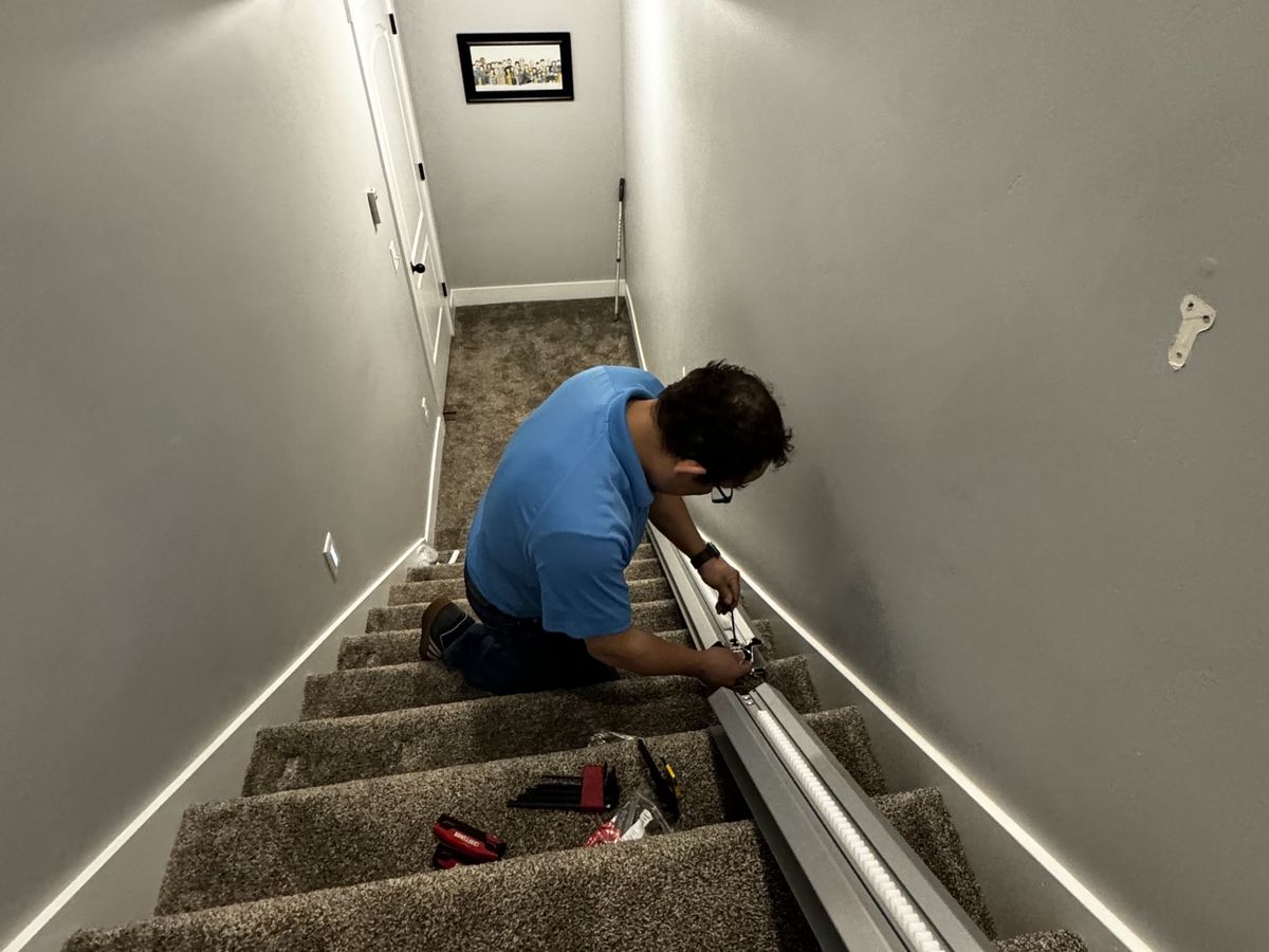 Installer kneeling on a carpeted basement staircase, fastening a stair lift rail to the steps