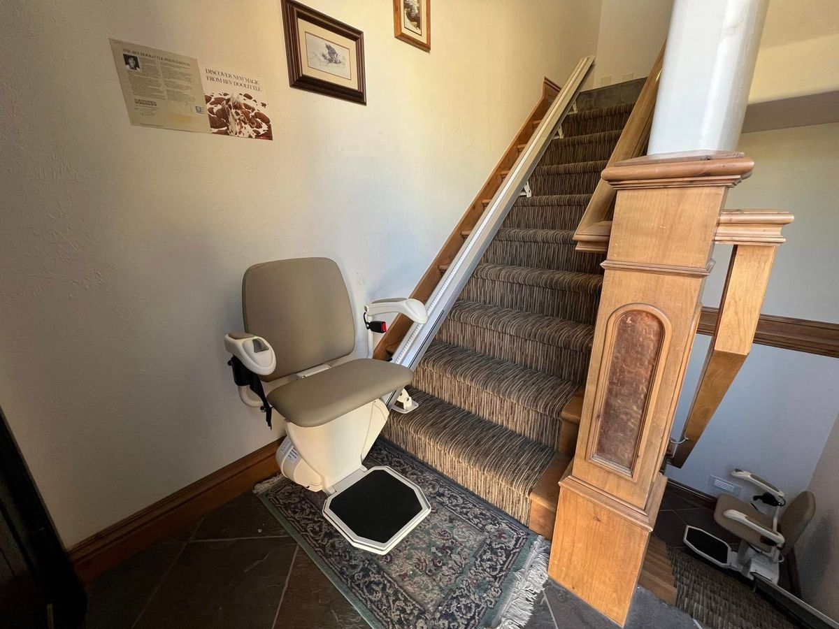 Lazo install: tan-seated lift at the foot of a runner staircase beside a carved newel post