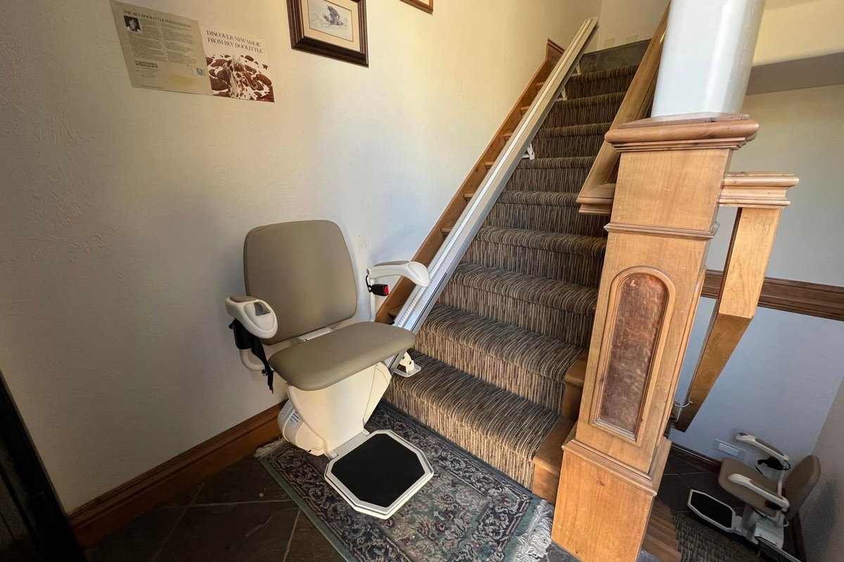 Harmar SL600 stair lift at the foot of a staircase in a tiled entry, rug and wood newel post beside it